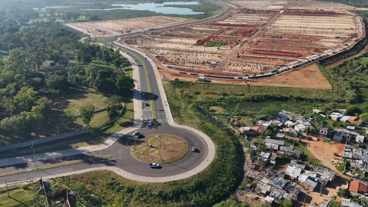 Vista aérea de una rotonda asfaltada junto a un amplio terreno nivelado para obras y un sector de viviendas precarias.