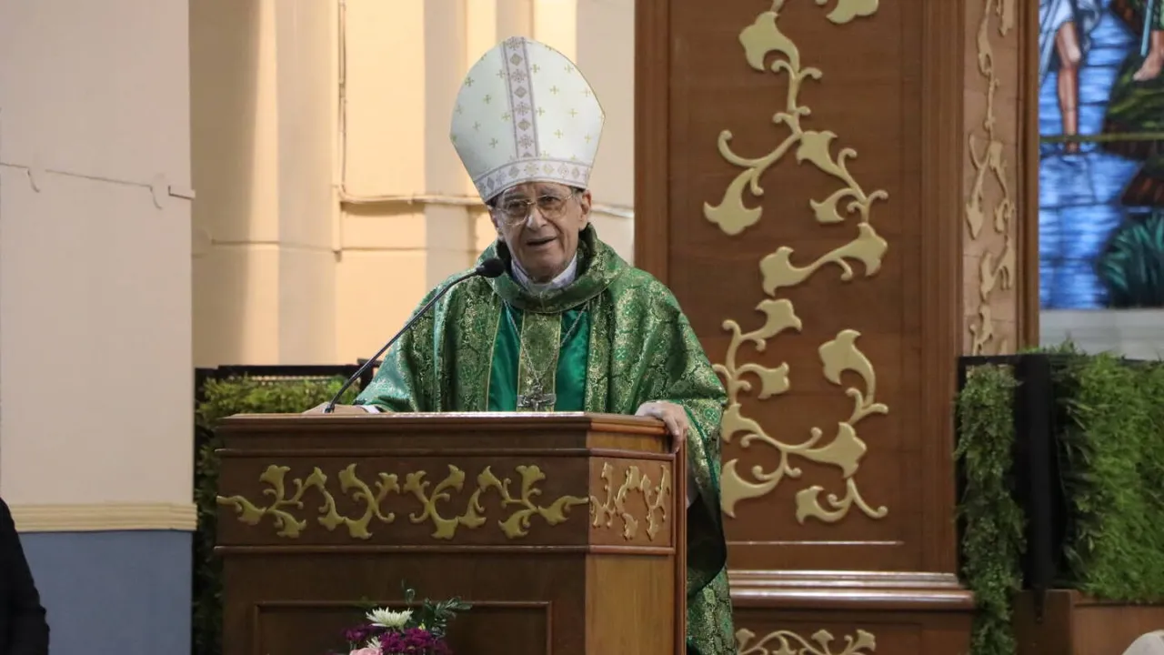Monseñor con mitra blanca y vestimenta litúrgica verde habla desde un púlpito de madera con micrófono.