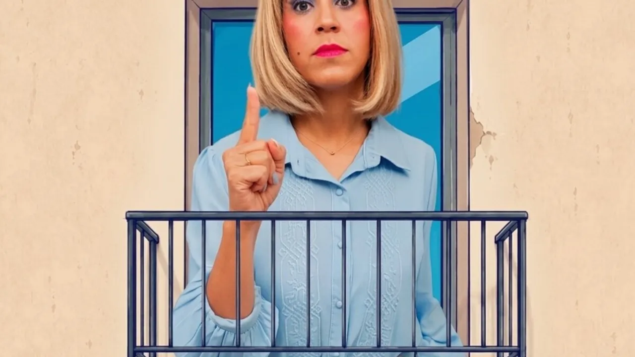 Mujer rubia con camisa celeste mirando al frente y levantando el dedo índice desde un balcón teatral.