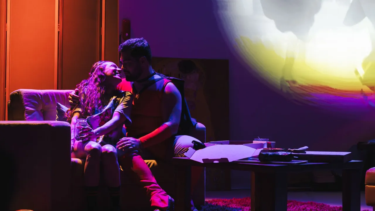 Dos actores en escena sentados en un sillón dialogando bajo una iluminación teatral de tonos morados y cálidos.