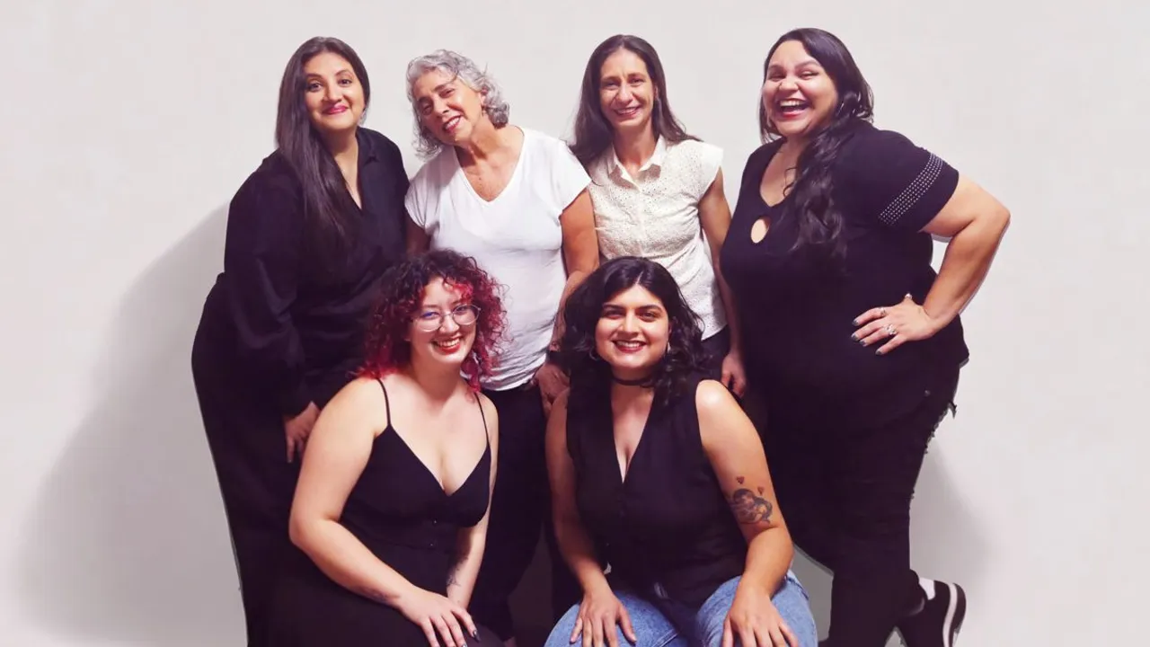 Seis mujeres sonríen juntas y posan de pie y agachadas frente a un fondo blanco claro en un estudio.