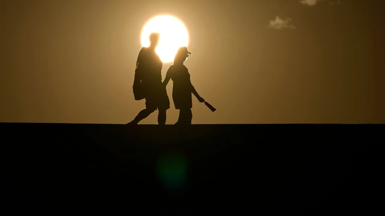 Siluetas de dos personas caminando con un bolso bajo un sol brillante y cielo despejado.
