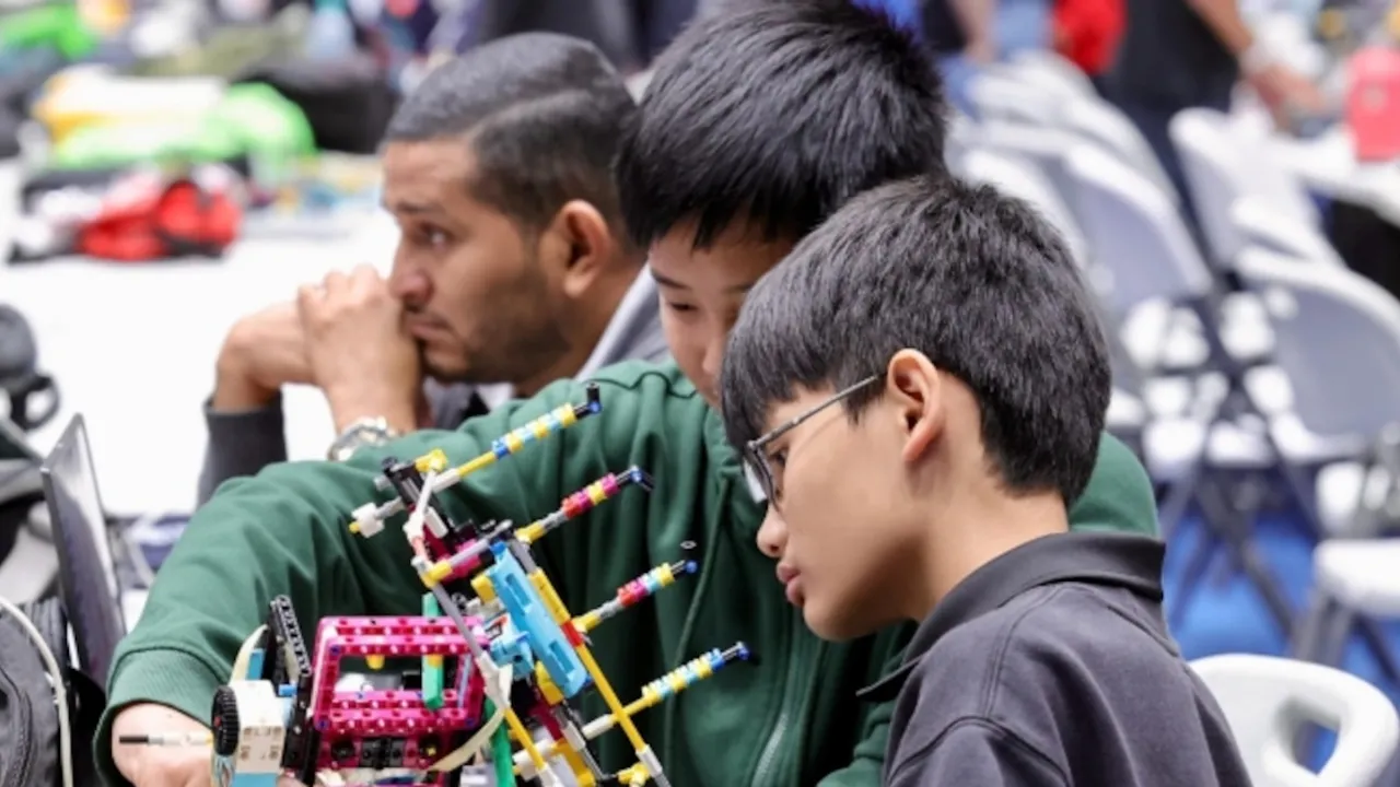 Dos jóvenes estudiantes concentrados arman una estructura robótica con piezas de plástico de colores.