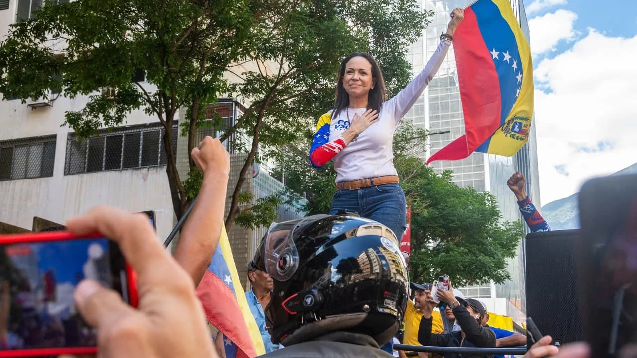 María Corina Machado saluda a una multitud y sostiene una bandera venezolana durante una concentración pública.
