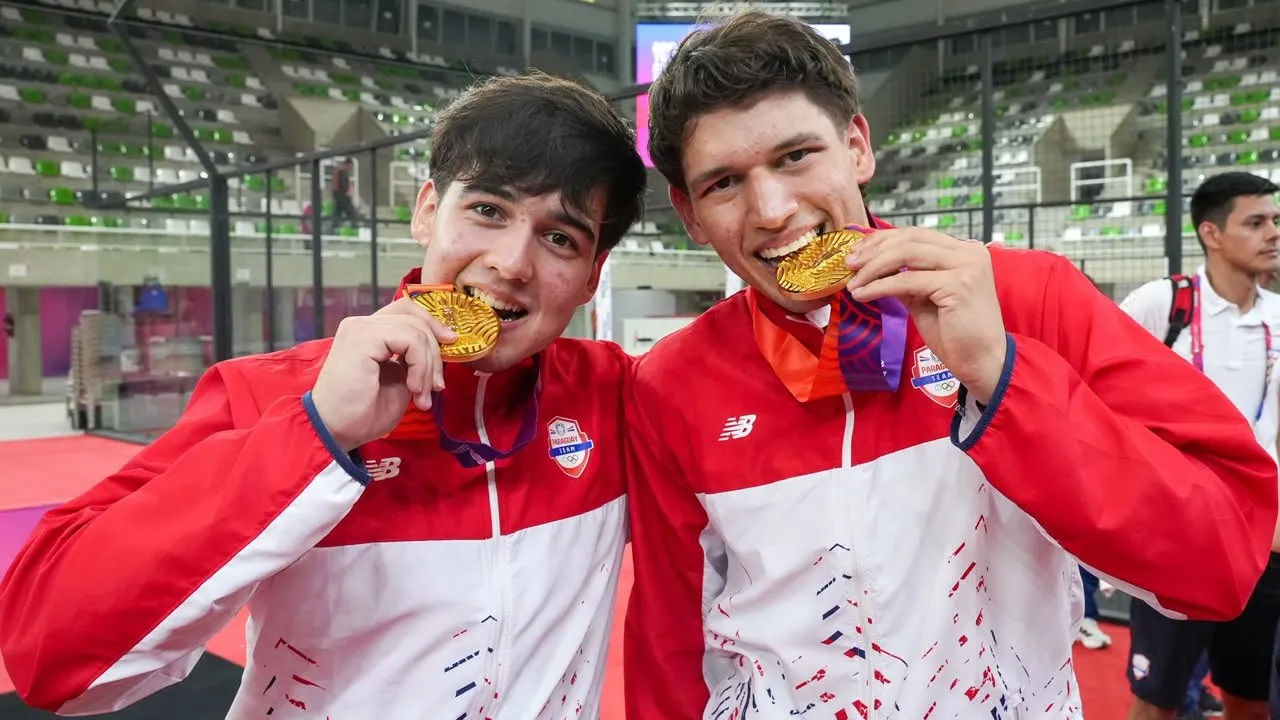 Dos deportistas con uniforme rojo y blanco muerden sonrientes sus medallas doradas en un estadio cubierto.