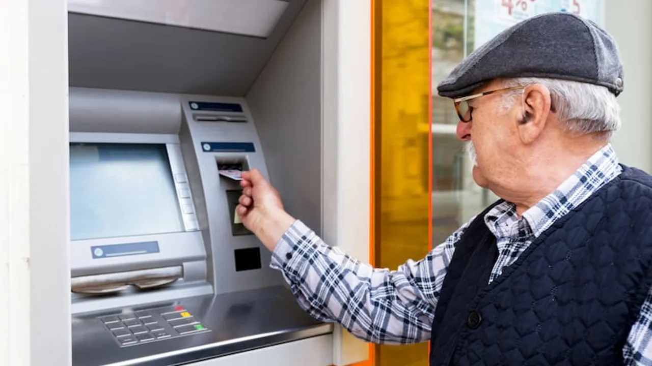 Hombre de edad avanzada con boina y chaleco introduciendo una tarjeta bancaria en un cajero automático en la calle