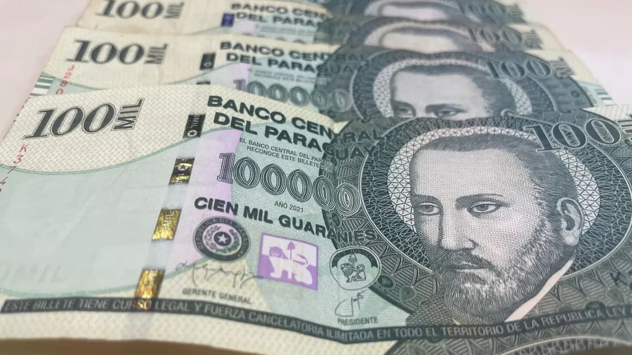 Varios billetes paraguayos de cien mil guaraníes dispuestos en fila sobre una superficie plana clara.