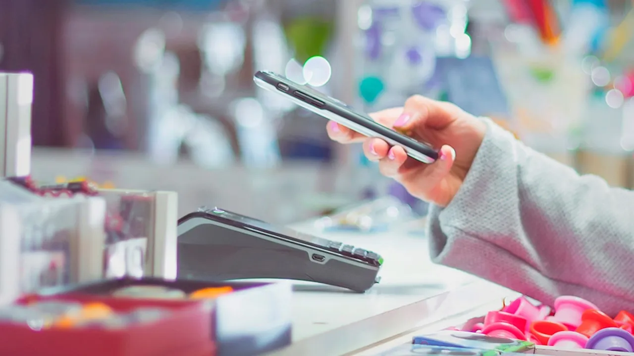 Mano de una persona sosteniendo un teléfono inteligente sobre un datáfono de cobro electrónico en una tienda comercial