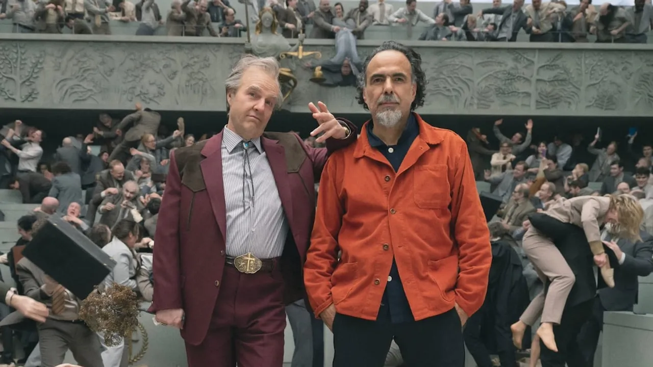 Dos hombres posan frente a una multitud caótica de fondo teatral, uno con traje bordo y otro con campera naranja.