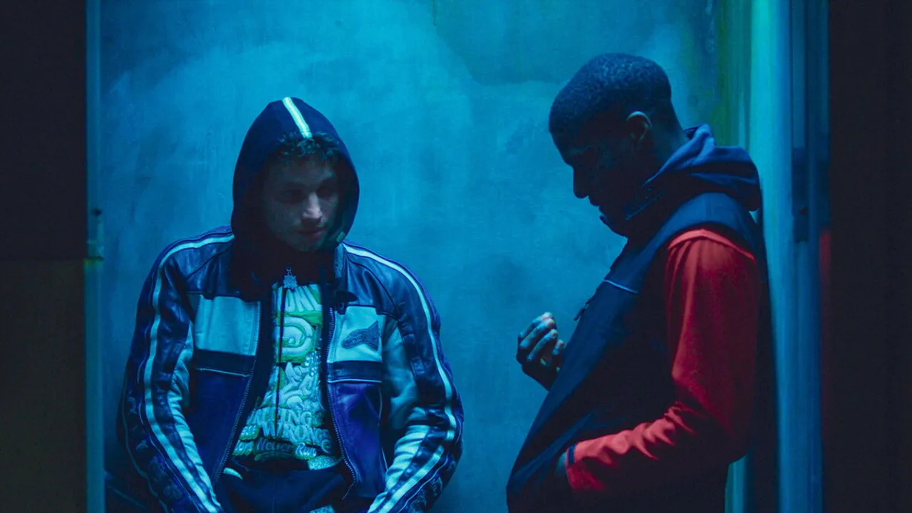Dos jóvenes dialogan bajo una iluminación azul tenue en una escena cinematográfica de suspenso urbano.