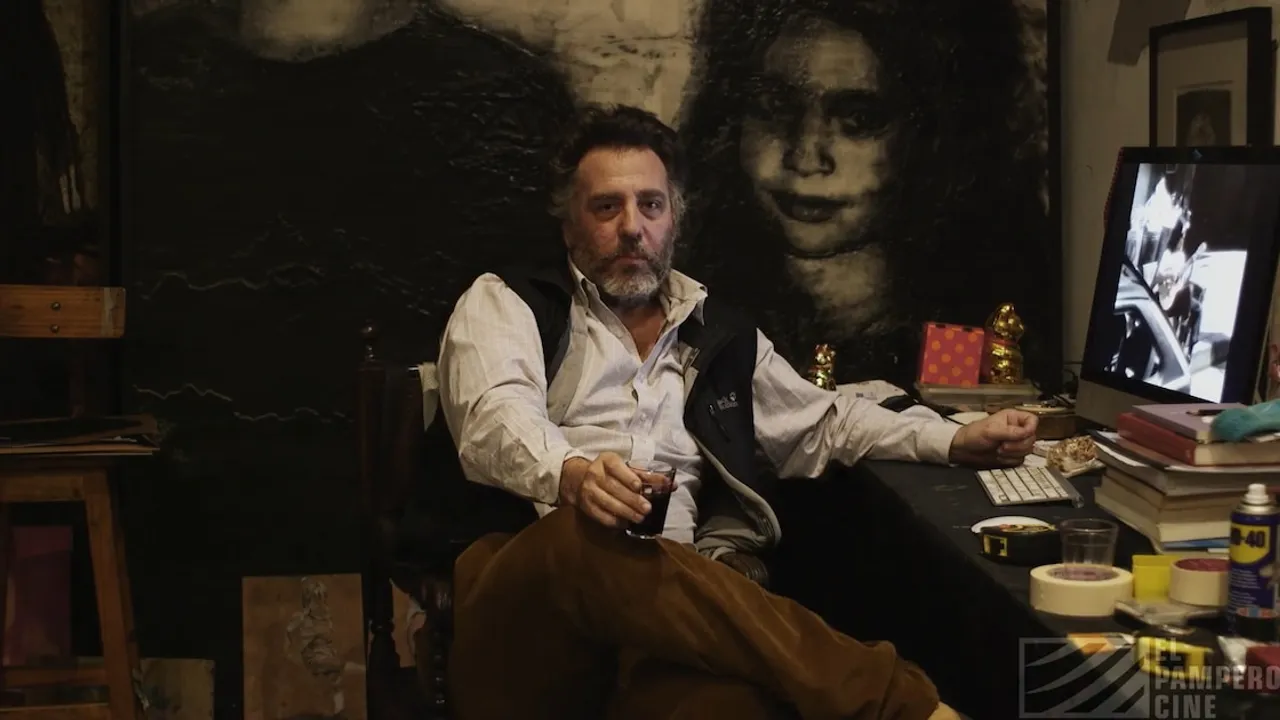 Hombre con barba sentado frente a un escritorio con un vaso en la mano, cerca de una pantalla y pinturas oscuras.