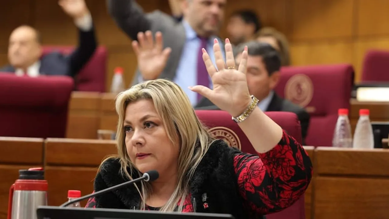 Una legisladora vestida de rojo y negro levanta la mano derecha durante una sesión parlamentaria.