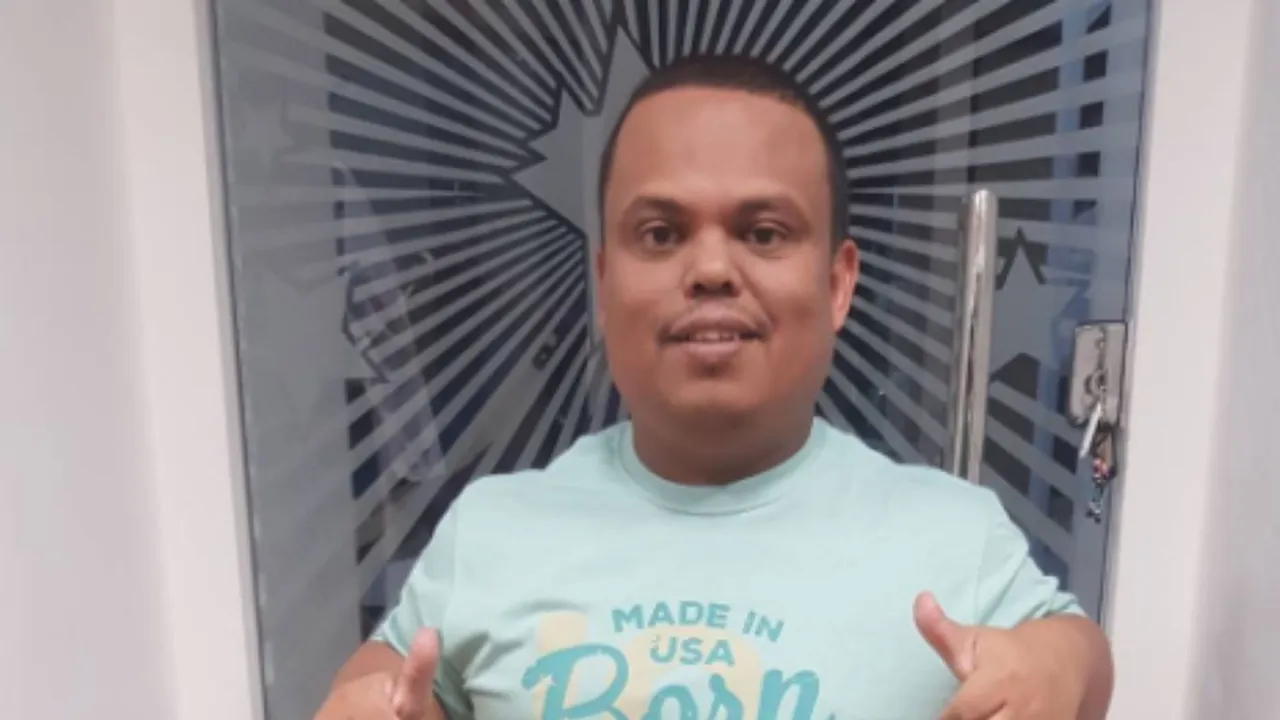Hombre de talla baja sonríe y hace un gesto positivo frente a una puerta de vidrio con diseño radial.