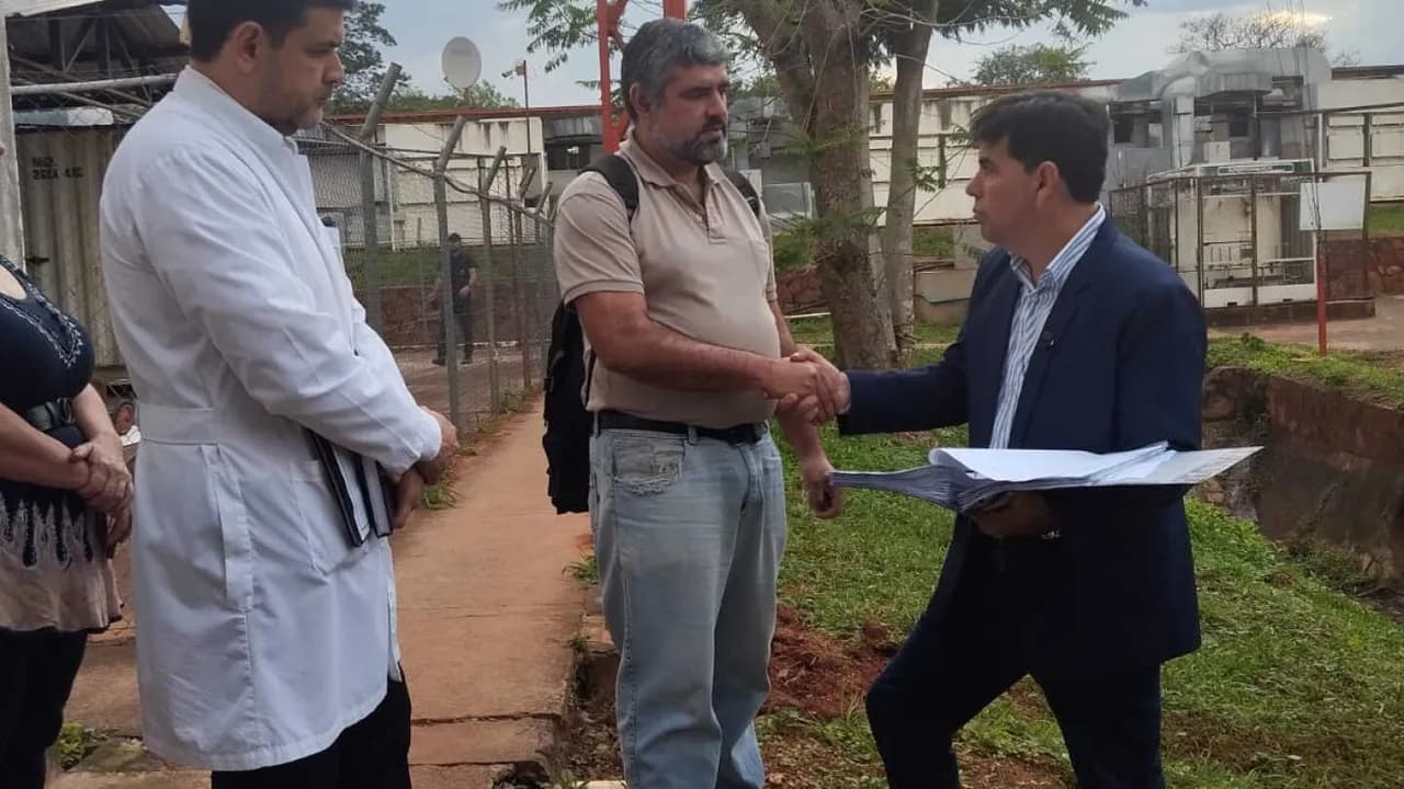 Un médico con bata blanca y un diputado de traje azul se dan la mano frente a la planta de oxígeno del hospital.