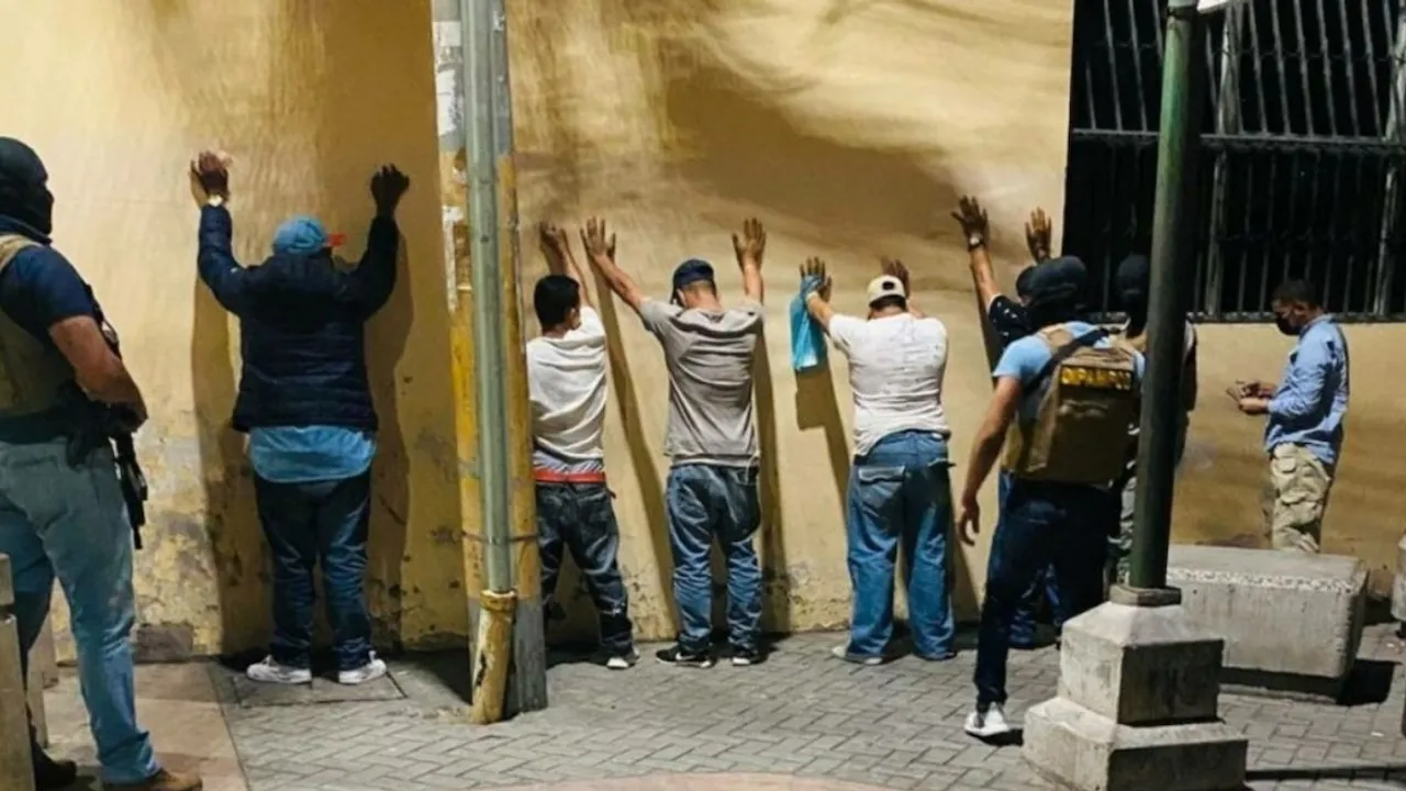 Agentes policiales armados custodian a varios sospechosos colocados de cara a una pared durante un operativo.