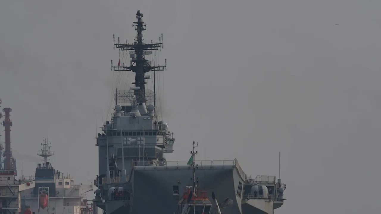 Estructura principal y torre de mando de un gran buque de guerra gris amarrado en un puerto comercial.