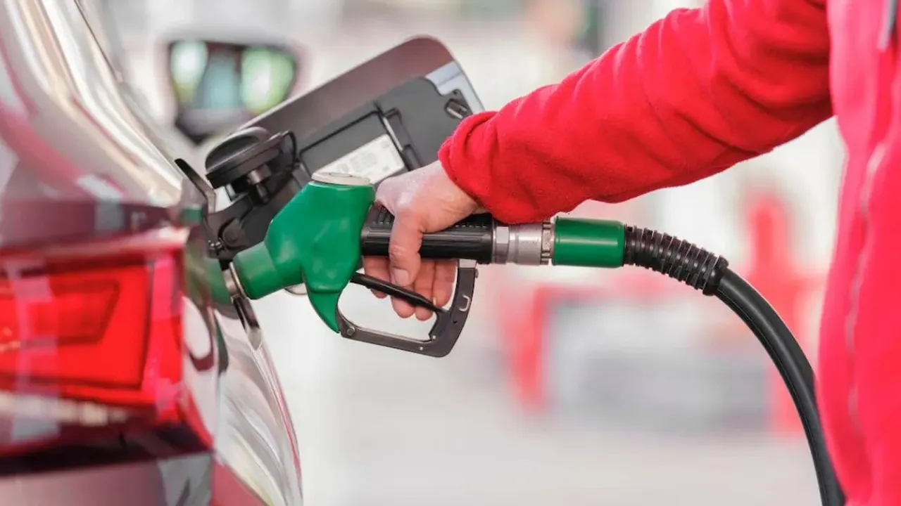 Una persona con campera roja sostiene la manguera con pico verde mientras carga combustible en un automóvil.
