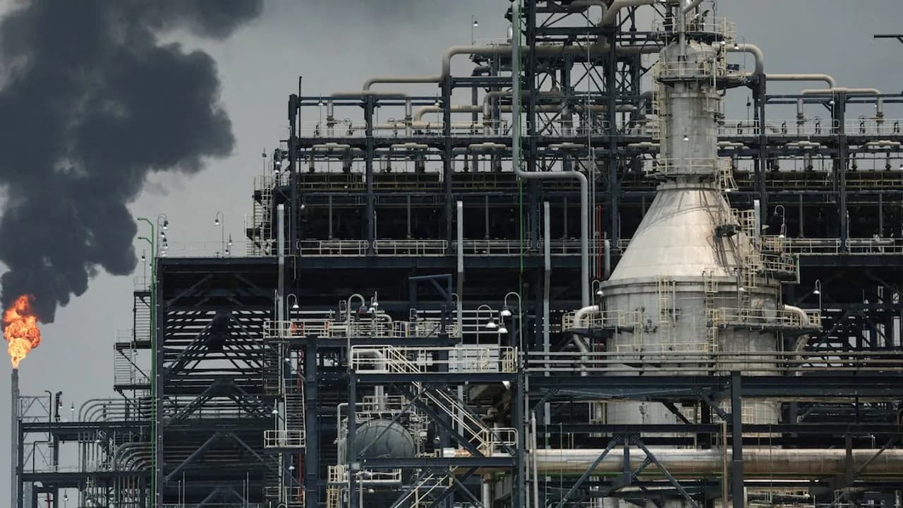 Estructuras metálicas de una refinería industrial con una chimenea expulsando fuego y humo oscuro.