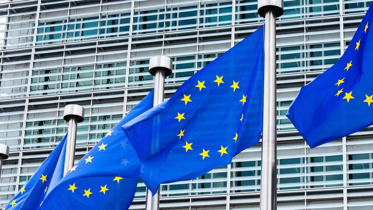 Banderas azules de la Unión Europea ondean en mástiles frente a la fachada acristalada de un edificio institucional.