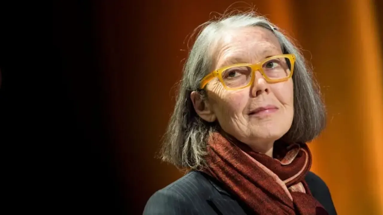 Anne Carson sonríe con anteojos amarillos y bufanda marrón frente a un fondo difuminado en penumbra.