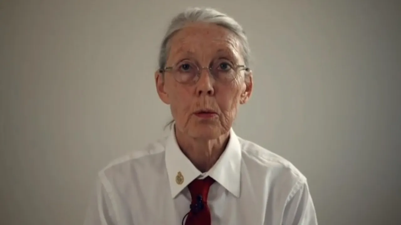 Retrato de Anne Carson con cabello gris, gafas redondas, camisa blanca con insignia en el cuello y corbata roja.