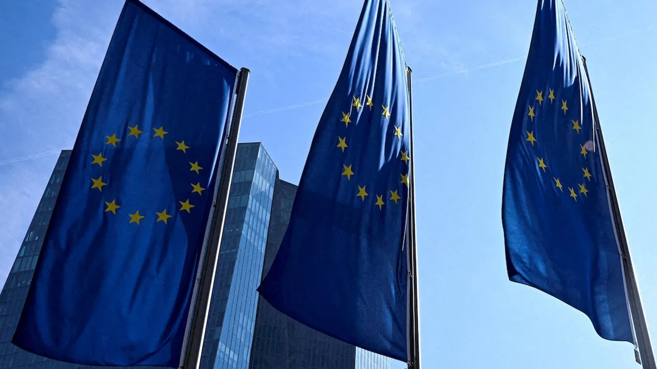 Tres banderas azules con las estrellas amarillas de la Unión Europea ondeando frente a un moderno edificio vidriado.