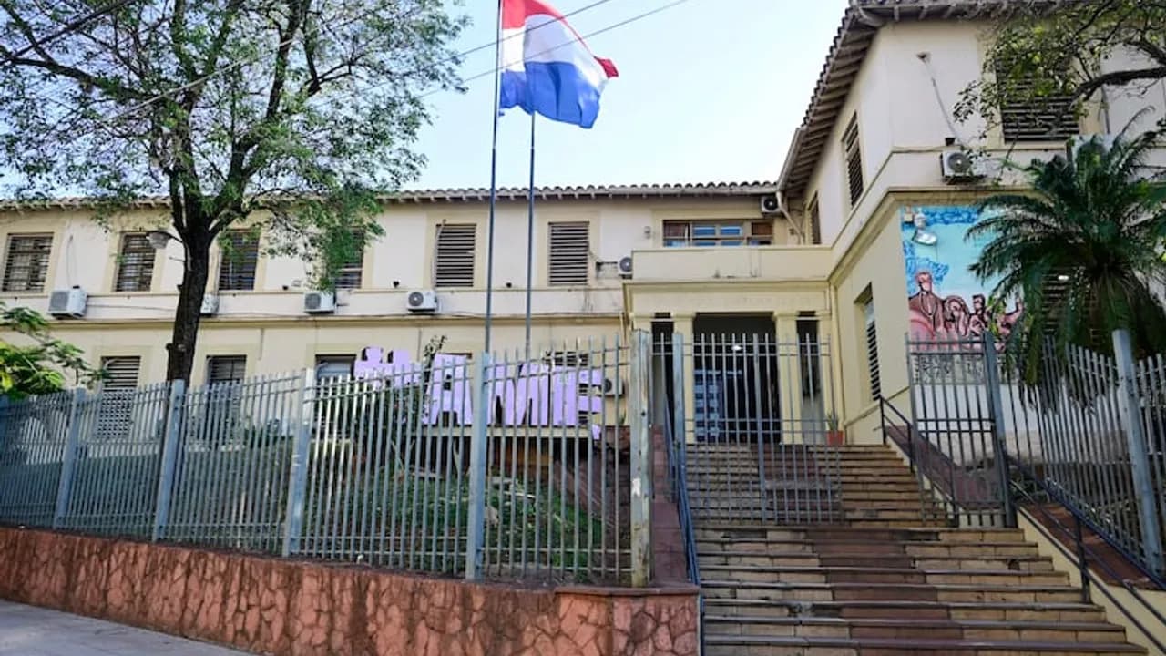 Fachada de un centro asistencial con rejas perimetrales, escalinata de acceso y la bandera de Paraguay flameando.