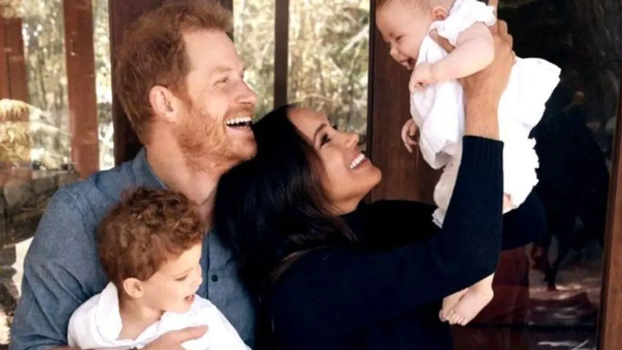 El príncipe Enrique y Meghan Markle sonríen en un entorno al aire libre mientras sostienen en brazos a sus dos pequeños hijos.
