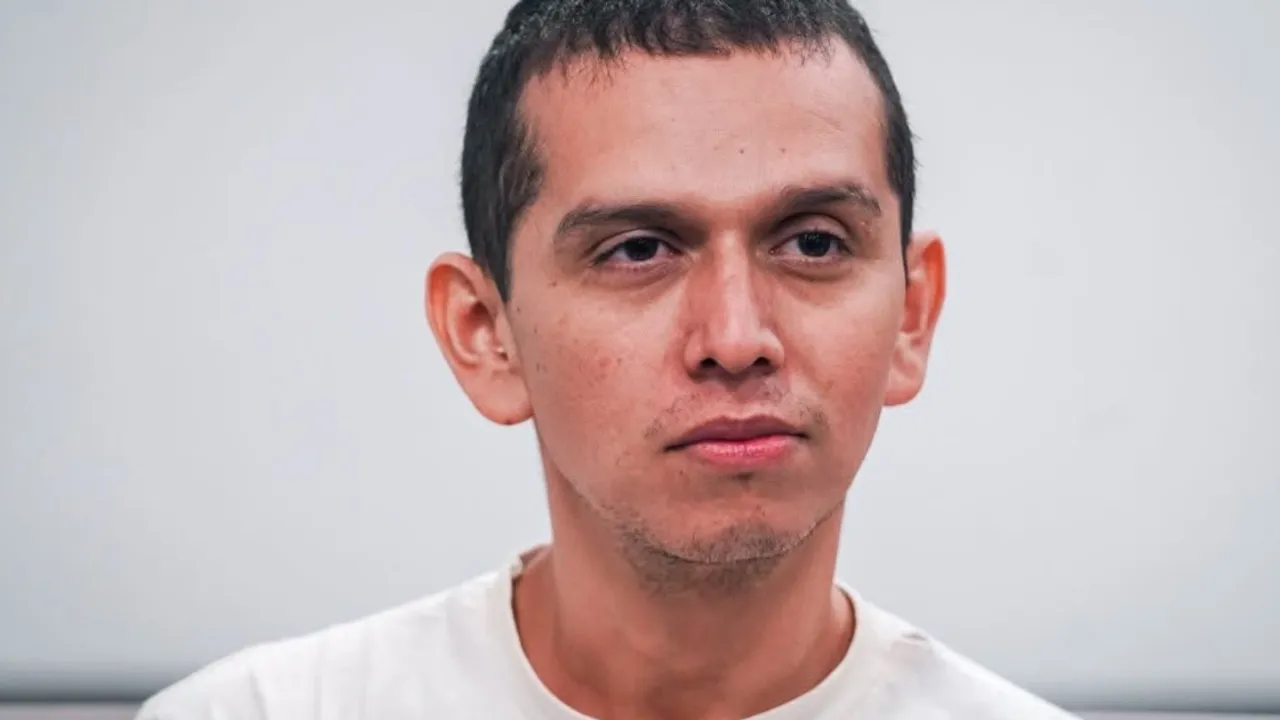 Hombre joven con cabello corto oscuro y camiseta blanca mira de frente con expresión seria.