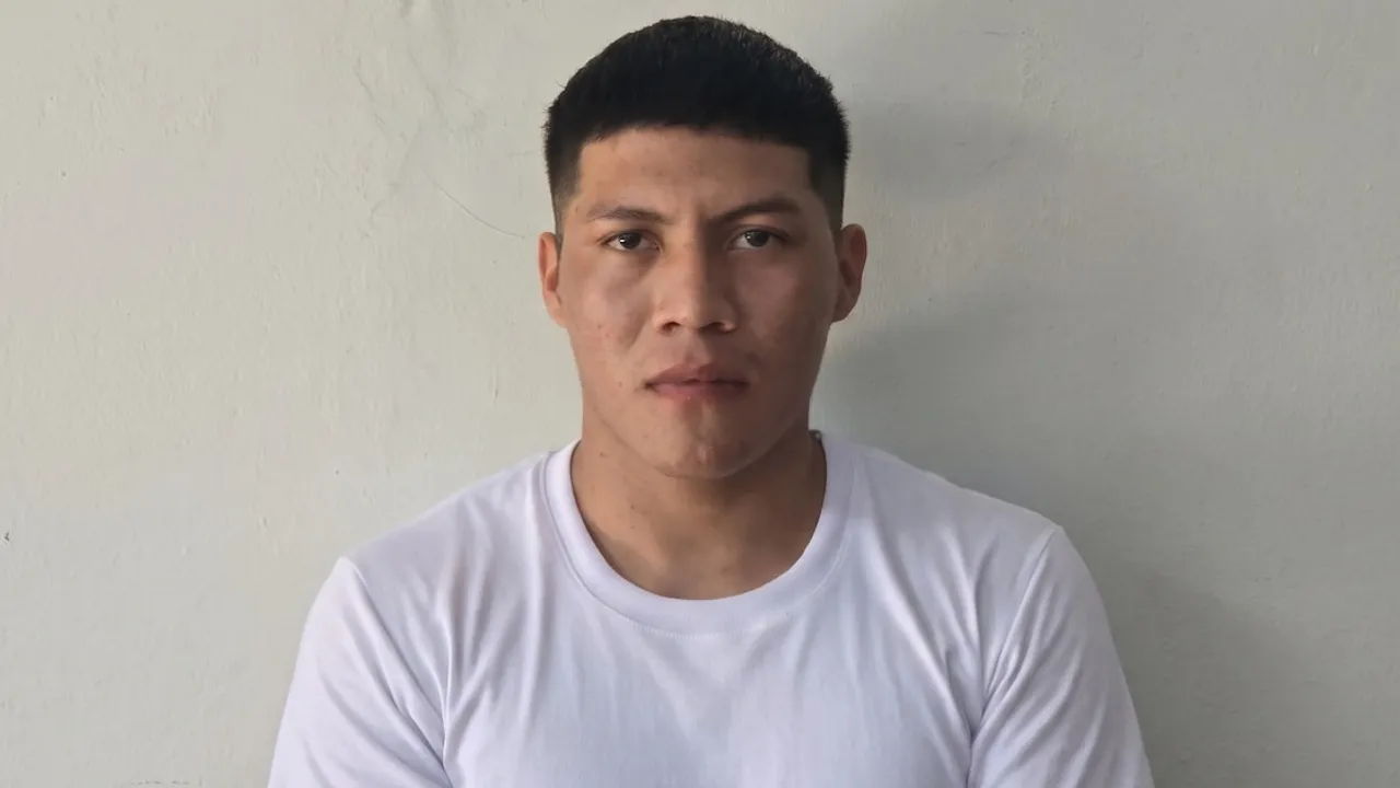 Hombre joven con cabello corto oscuro y camiseta blanca mira fijamente hacia el frente ante una pared clara