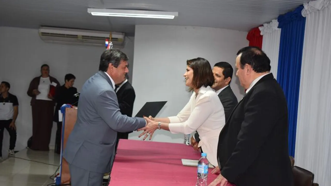 Autoridades entregan un certificado a un hombre durante un acto protocolar frente a una mesa oficial.