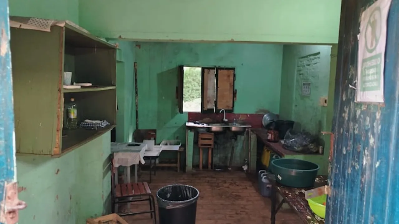 Espacio precario utilizado como cocina y comedor escolar con paredes verdes, mesada con bacha y utensilios plásticos.