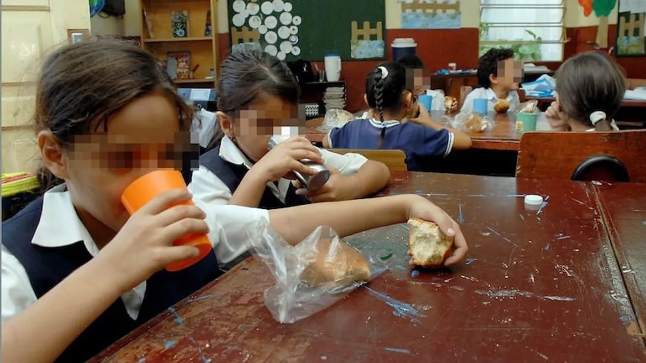Niños en un aula escolar sentados en mesas de madera mientras toman bebidas y comen pan durante el recreo.