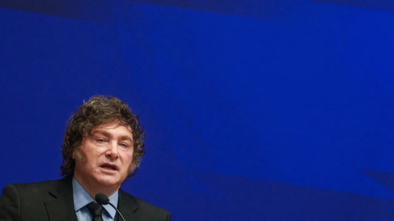 Javier Milei habla frente a un micrófono con traje oscuro sobre un fondo azul durante una intervención.