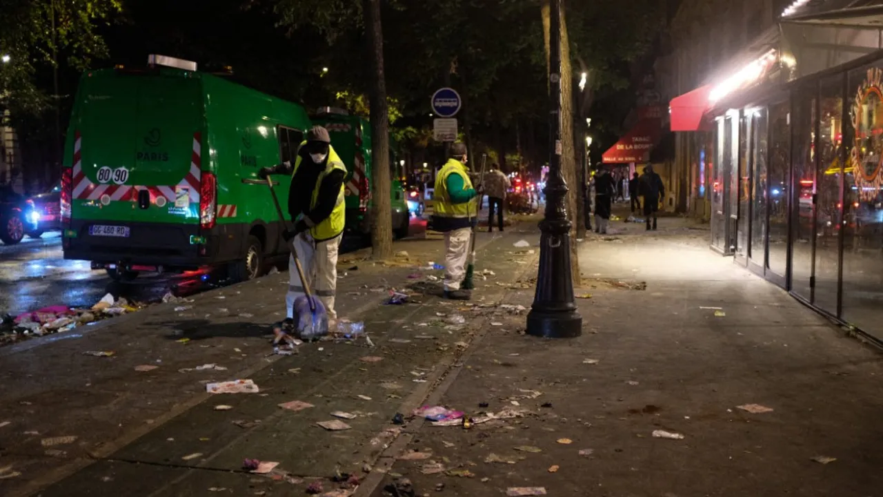 Trabajadores de limpieza barren restos y basura en una vereda nocturna junto a furgonetas municipales de París.
