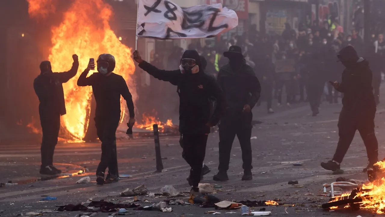 Manifestantes con rostros cubiertos caminan por una calle junto a barricadas en llamas y humo denso.