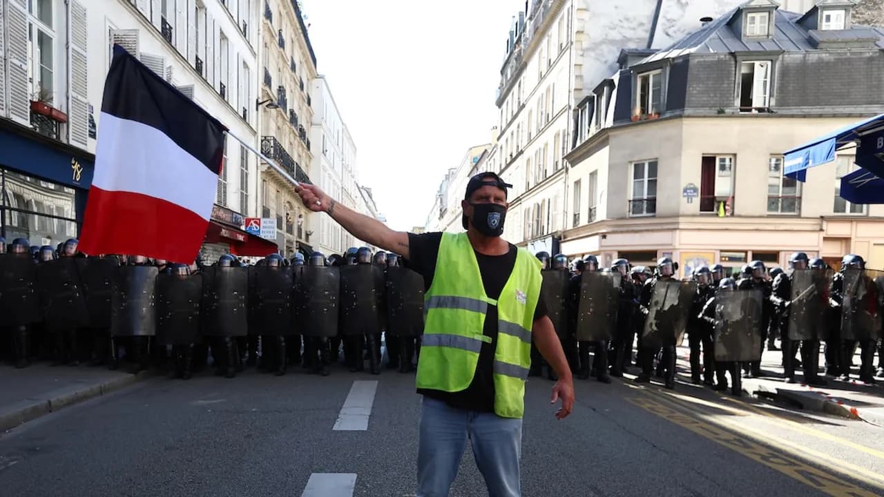 Hombre con chaleco reflectante y mascarilla ondea la bandera de Francia frente a una fila de policías antidisturbios.