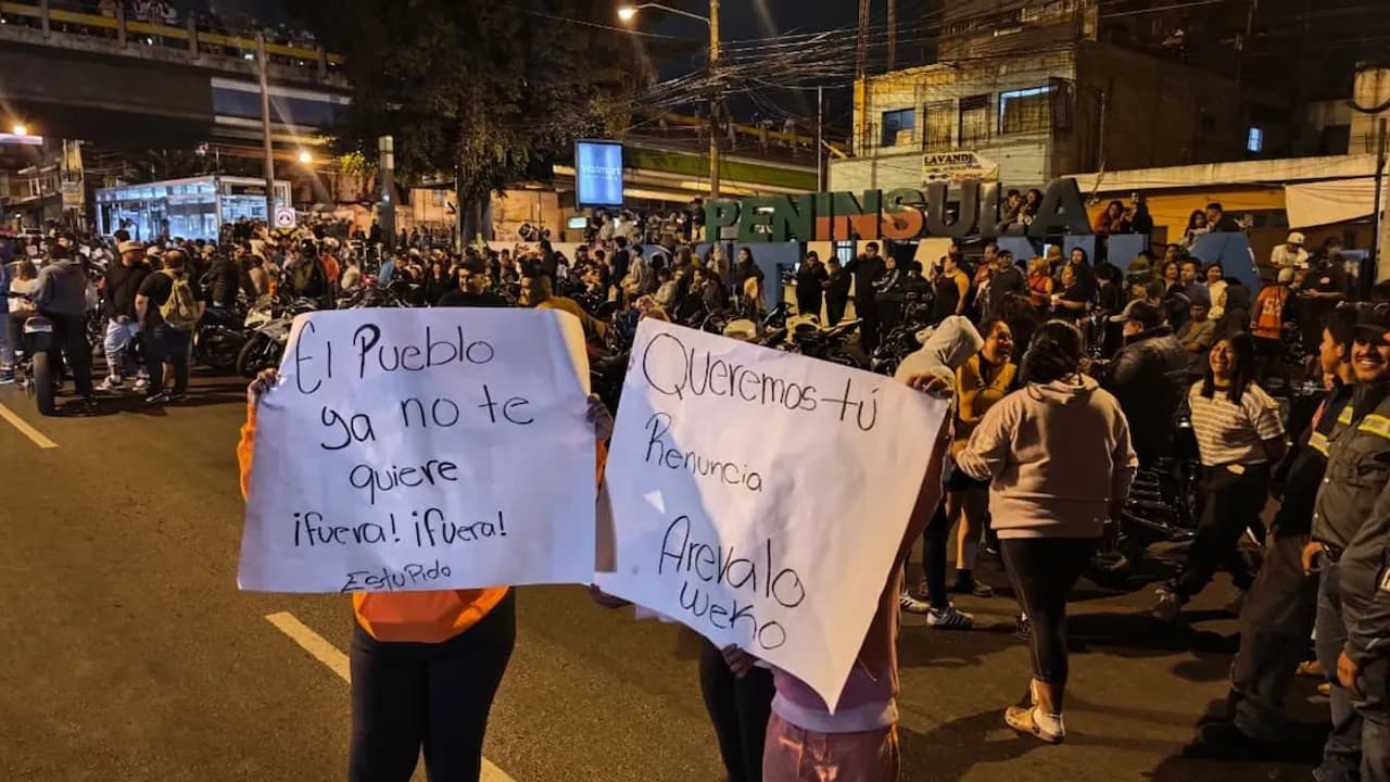 Personas sostienen carteles de protesta exigiendo la renuncia presidencial durante una manifestación nocturna en una avenida.