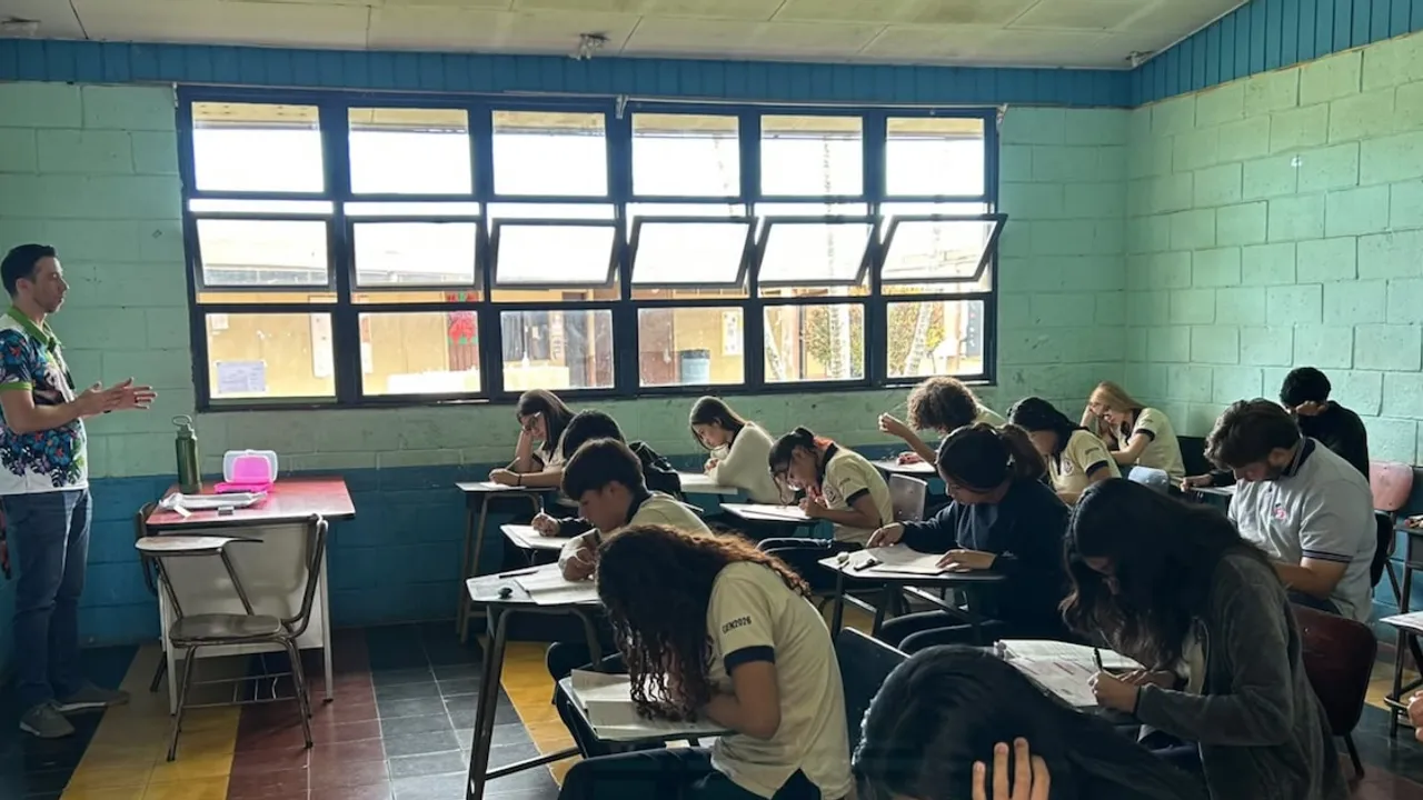 Alumnos sentados en pupitres rindiendo un examen dentro de un aula escolar frente a un docente.