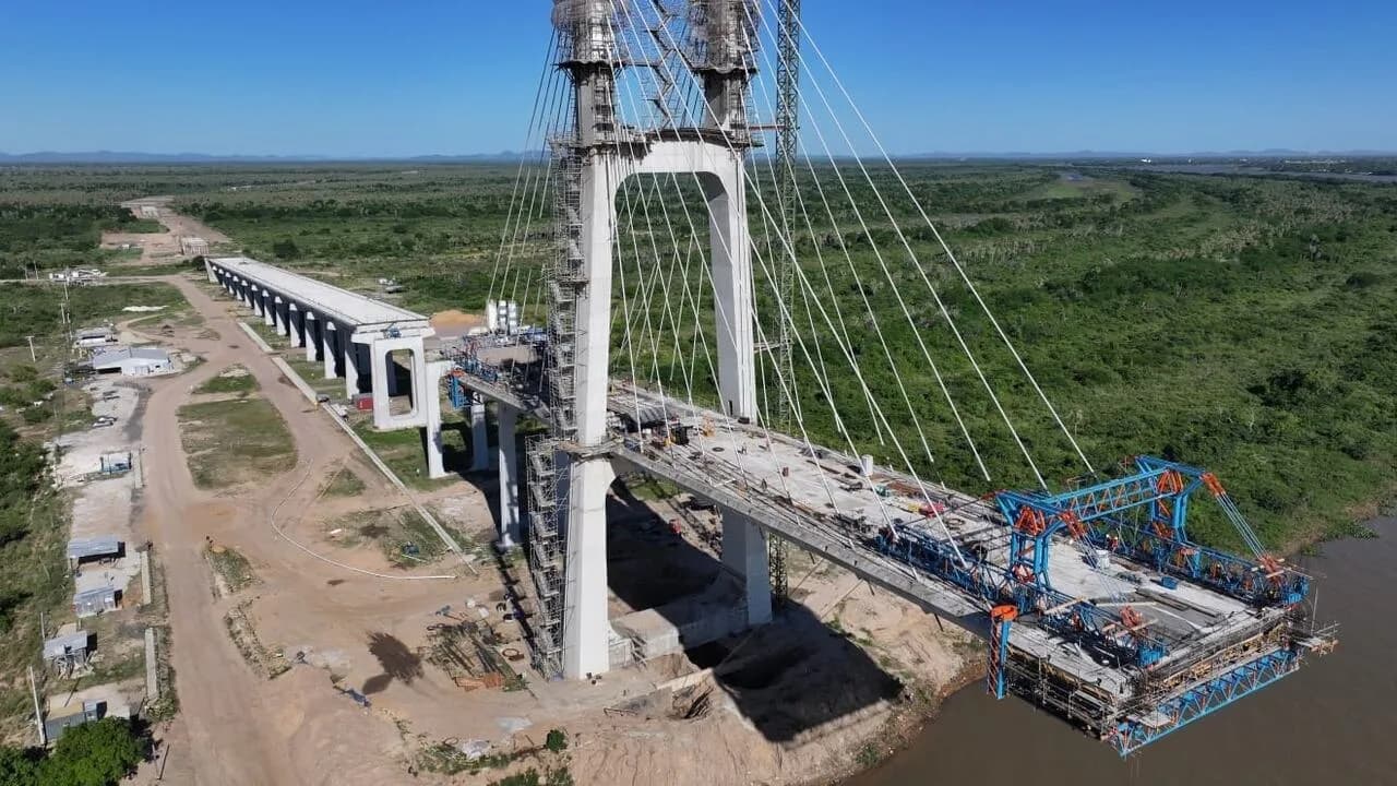 Estructura en construcción de puente atirantado con torre de hormigón blanco y grúas sobre el río Paraguay.
