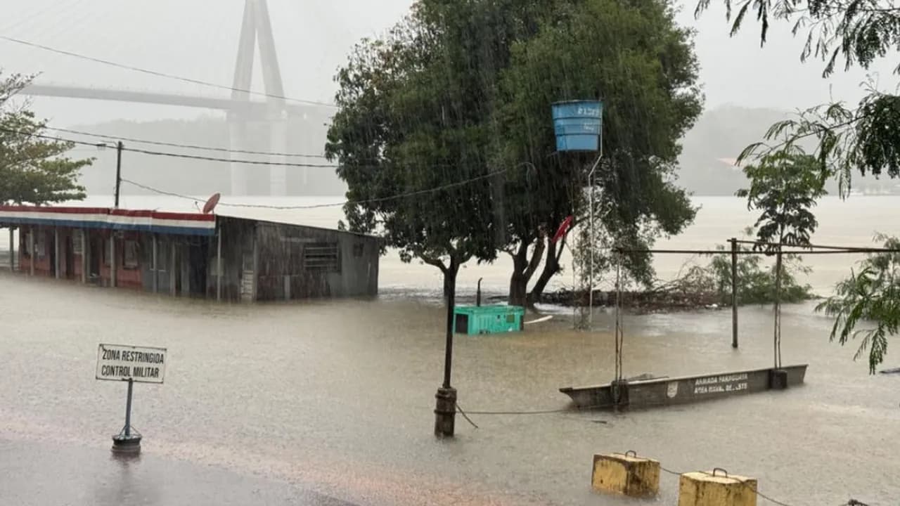 Caseta y orilla costera anegadas por la crecida del río junto a una embarcación de la Armada Paraguaya bajo la lluvia.