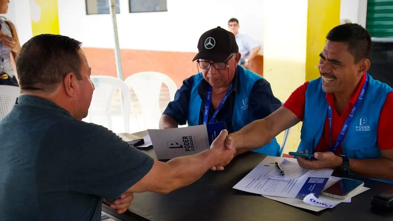 Tres personas sentadas frente a una mesa firman documentos y estrechan sus manos durante una actividad cívica.