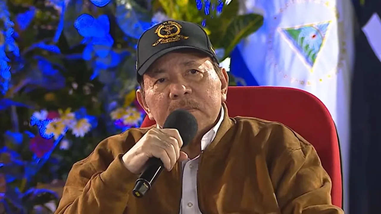 Daniel Ortega habla frente a un micrófono luciendo una gorra oscura y una campera marrón ante banderas oficiales.