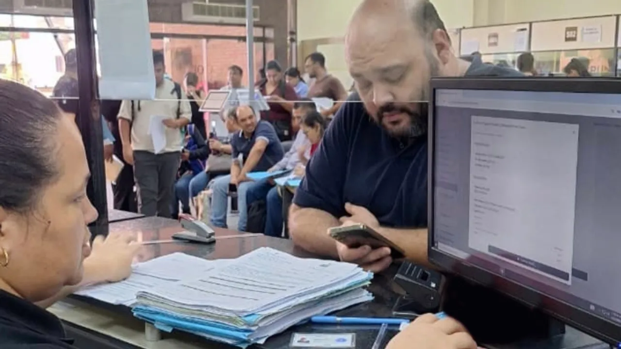 Un hombre observa su celular ante un mostrador de atención al público con expedientes impresos y computadoras.