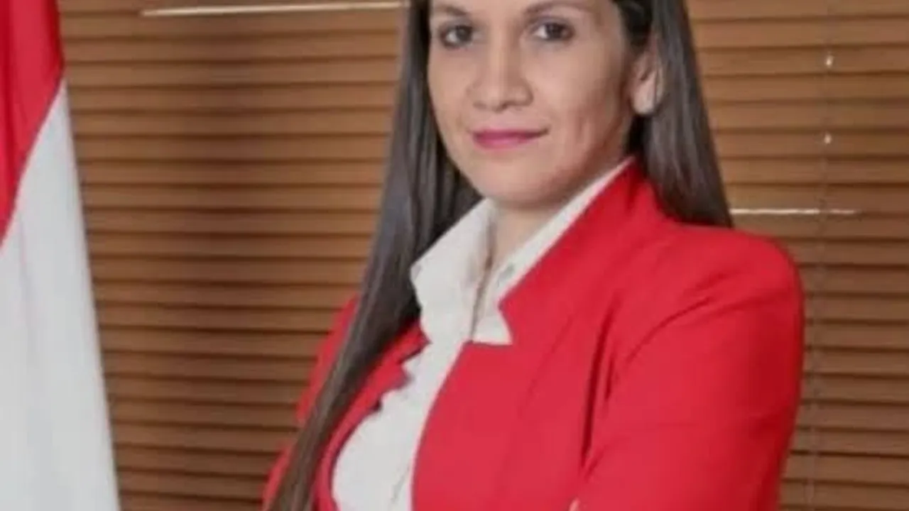 Carmen Marín vestida con saco rojo y camisa blanca mirando al frente junto a una bandera de Paraguay.