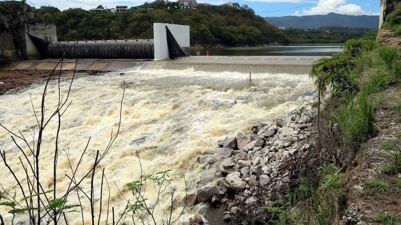 Agua correntosa fluyendo con fuerza a través del vertedero de una presa de concreto junto a rocas y colinas.
