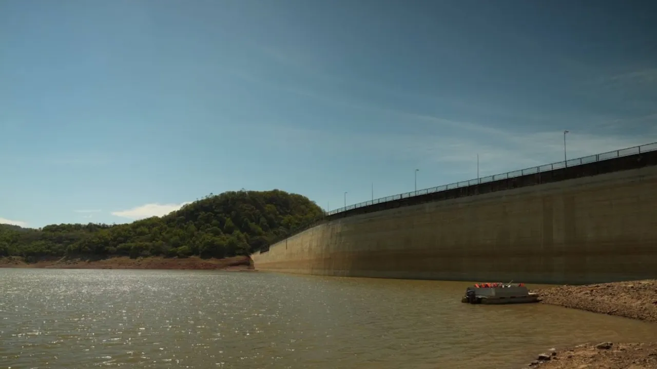 Muro de concreto de un embalse que retiene agua frente a una colina verde bajo el cielo despejado.