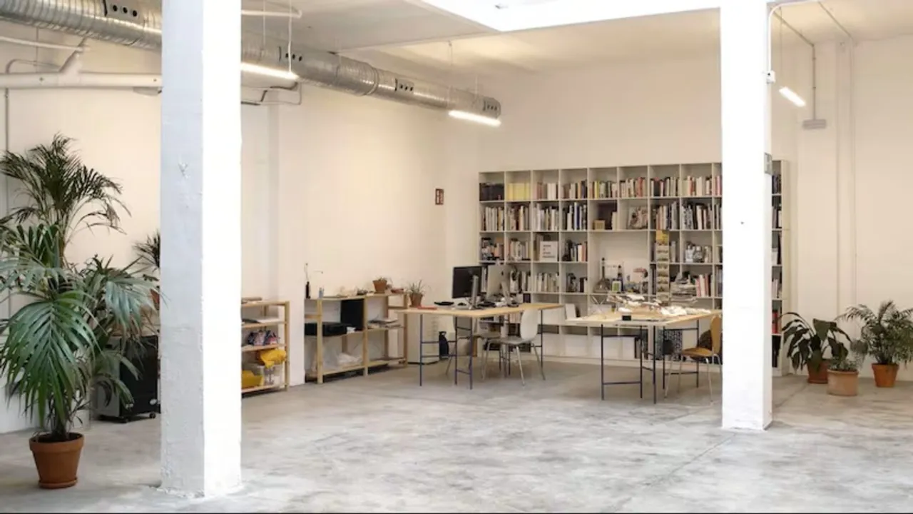 Amplio espacio de taller con columnas blancas, estanterías de libros, plantas en macetas y mesas de trabajo.