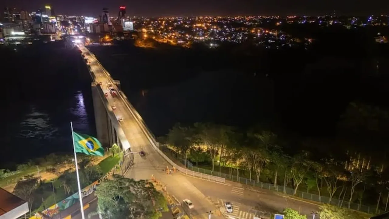 Vista aérea nocturna del Puente de la Amistad iluminado con la bandera de Brasil en primer plano.