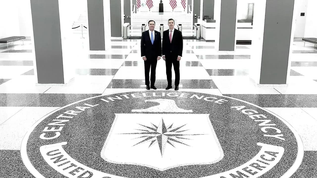 Dos hombres de traje posan de pie sobre el escudo circular de la CIA en el suelo de un amplio salón.